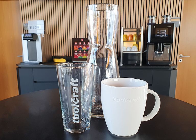 Glaskaraffe, Trinkglas und Kaffeebecher vor einer Kaffeemaschine und einem Wasserspender